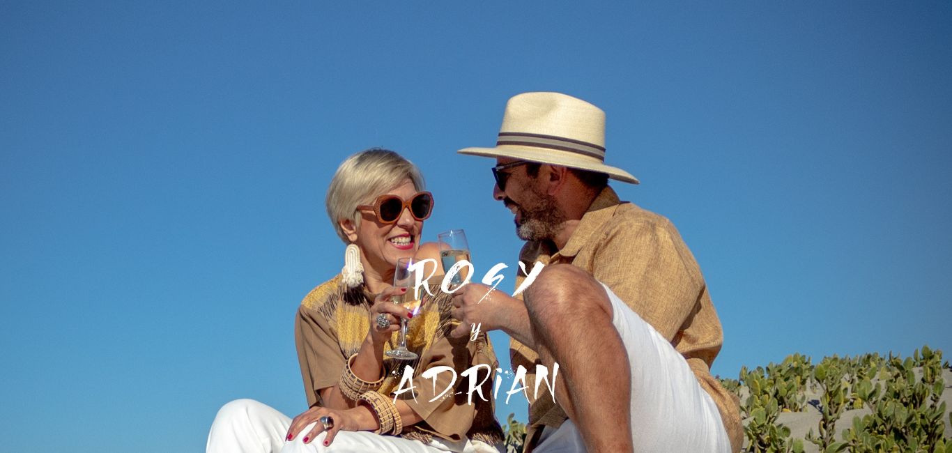 Rosy y Adrián sosteniendo una copa de espumoso, sentados en la arena con el cielo azul de fondo